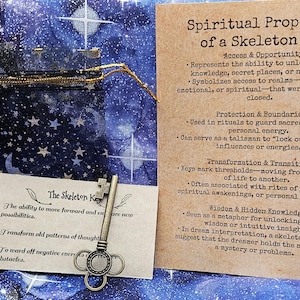 May include: A silver skeleton key rests on a card with text about its spiritual properties. The key is next to a small card with text about the key's ability to move forward. The background is a blue fabric with a celestial pattern.