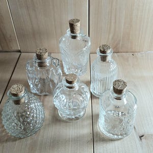 May include: A collection of six clear glass bottles of varying shapes and textures, each topped with a cylindrical cork stopper. The bottles feature distinct embossed patterns including diamond, vertical ribbing, and floral motifs.