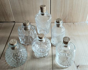 May include: A collection of six clear glass bottles of varying shapes and textures, each topped with a cylindrical cork stopper. The bottles feature distinct embossed patterns including diamond, vertical ribbing, and floral motifs.