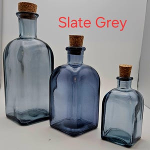 May include: Three slate grey glass bottles with cork stoppers. The bottles vary in size, with a rectangular shape and rounded shoulders. The text "Slate Grey" is displayed in red. The bottles are empty and sit on a white surface.