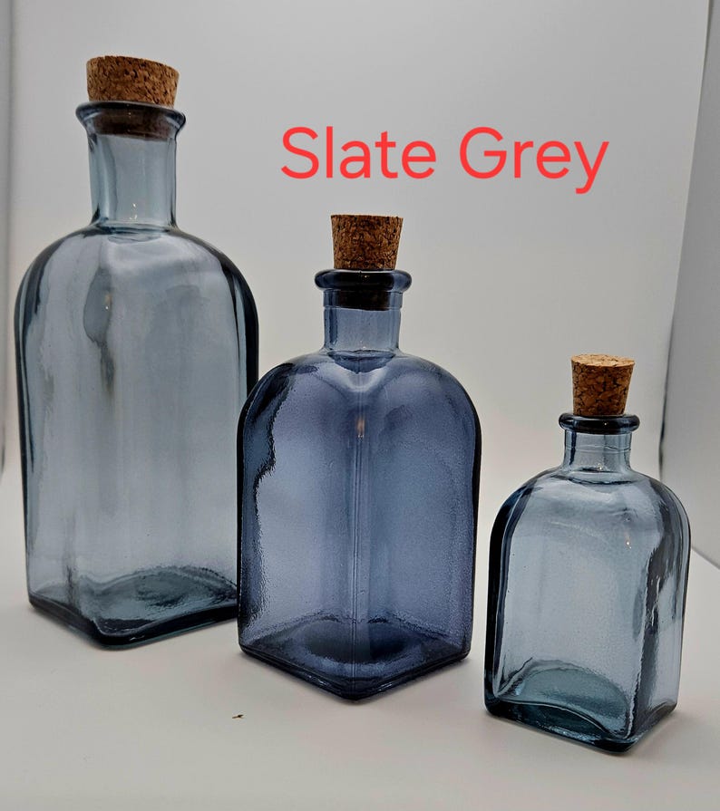 May include: Three slate grey glass bottles with cork stoppers. The bottles vary in size, with a rectangular shape and rounded shoulders. The text "Slate Grey" is displayed in red. The bottles are empty and sit on a white surface.