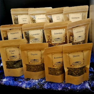 May include: A collection of twelve small, light brown paper pouches with clear windows, each containing dried herbs or tea leaves. Labels on the pouches feature the names Lemon Balm, Chamomile, Holy Basil, Echinacea, Peppermint, Lavender, Calendula, Lemongrass, Green Tea, Thyme, Rosemary, and Stevia.