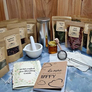 May include: A collection of dried herbs in brown pouches labeled Nettle Leaf, Howthorn Berries, Spruce Tips, Rose Petals, White Pine Needles, and Mullein, with a metal canister, white mortar and pestle, honey jar, tea strainer, and two booklets.