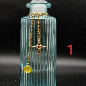 May include: A light blue, vertically ribbed glass bottle with a clear, faceted stopper. A gold-toned chain is draped around the neck, featuring a four-pointed star charm with a central stone and a small, round, translucent yellow bead pendant. The number 1 is visible.