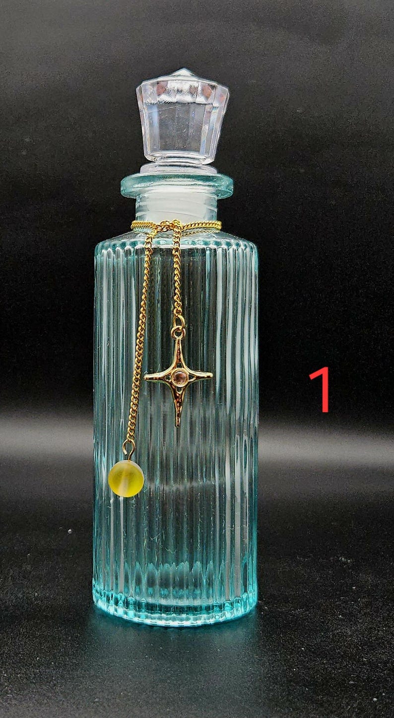 May include: A light blue, vertically ribbed glass bottle with a clear, faceted stopper. A gold-toned chain is draped around the neck, featuring a four-pointed star charm with a central stone and a small, round, translucent yellow bead pendant. The number 1 is visible.