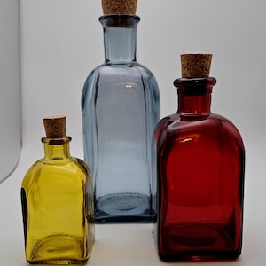 May include: Three coloured glass bottles with cork stoppers. The bottles are yellow, blue, and red. They have a rectangular shape with rounded edges. The blue bottle is the tallest, with the others being smaller in size.