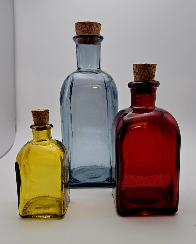 May include: Three coloured glass bottles with cork stoppers. The bottles are yellow, blue, and red. They have a rectangular shape with rounded edges. The blue bottle is the tallest, with the others being smaller in size.