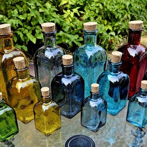 May include: A selection of coloured glass bottles with cork stoppers. The bottles are in shades of green, yellow, blue, and red. They are arranged on a glass surface, with foliage visible in the background. Measurements are not available.