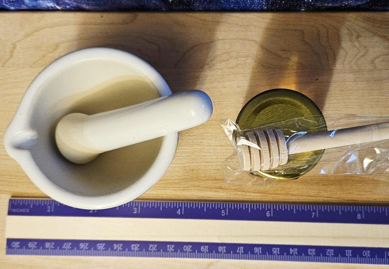 Może przedstawiać: Biały ceramiczny moździerz z tłuczkiem obok drewnianej łyżki do miodu owiniętej w przezroczystą folię, na jasnej drewnianej powierzchni. Fioletowa linijka na dole pokazuje wymiary w centymetrach do 30 cm.
