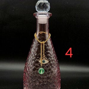 May include: A pink glass bottle with a textured honeycomb pattern, topped with a clear faceted crystal stopper. A gold-toned chain hangs around the neck, featuring an eye-shaped charm with a blue center and a small green bead. The number 4 is visible.