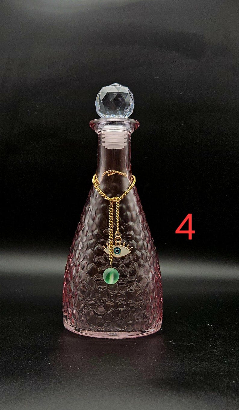 May include: A pink glass bottle with a textured honeycomb pattern, topped with a clear faceted crystal stopper. A gold-toned chain hangs around the neck, featuring an eye-shaped charm with a blue center and a small green bead. The number 4 is visible.