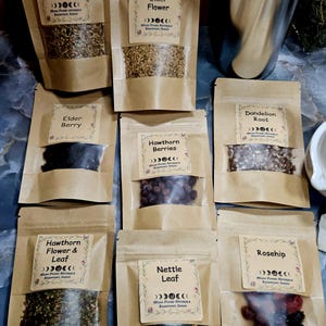 Może przedstawiać: Osiem brązowych papierowych torebek z suszonymi ziołami i jagodami, każda z etykietą Moon Phase Naturals, Sandpoint, Idaho. Etykiety identyfikują Yarrow Flowers, Elder Flower, Elder Berry, Hawthorn Berries, Dandelion Root, Hawthorn Flower & Leaf, Nettle Leaf oraz Rosehip.