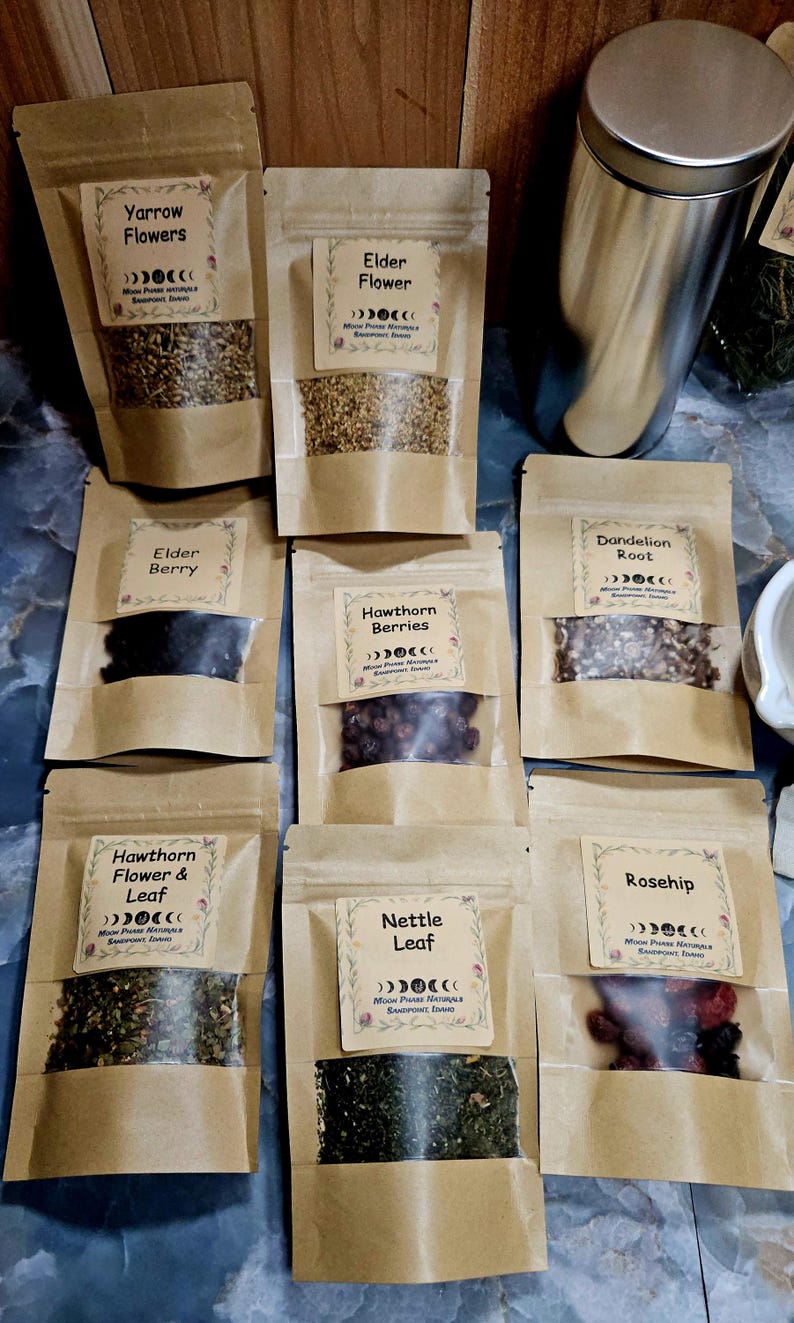 Może przedstawiać: Osiem brązowych papierowych torebek z suszonymi ziołami i jagodami, każda z etykietą Moon Phase Naturals, Sandpoint, Idaho. Etykiety identyfikują Yarrow Flowers, Elder Flower, Elder Berry, Hawthorn Berries, Dandelion Root, Hawthorn Flower & Leaf, Nettle Leaf oraz Rosehip.