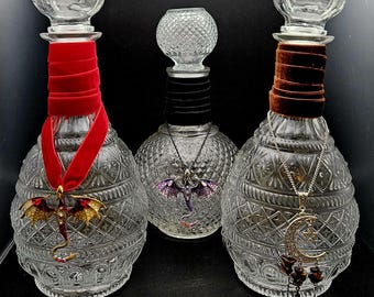 May include: Three decorative glass bottles with ornate diamond patterns. Each bottle has a different colored velvet ribbon or collar. One has a red ribbon with a dragon pendant, another a black collar with a dragon pendant, and the last a brown collar with a moon and star pendant.