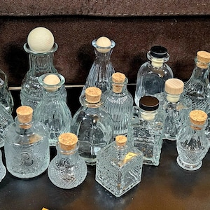 May include: Collection of clear glass bottles with cork and black stoppers. The bottles vary in shape and size, some with textured designs. A measuring tape is visible in the background.