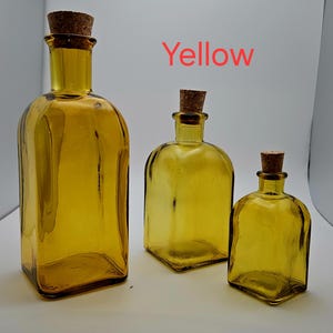 May include: Three yellow glass bottles with cork stoppers. The bottles vary in size, with a rectangular bottle on the left and two rounded square bottles to the right. The word "Yellow" is written in red above the bottles.