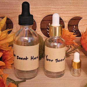 May include: Three clear glass bottles with black and gold droppers and labels that read "Your Scent Here" and "Your Scent". The bottles are surrounded by orange flowers and greenery, with a wooden background.