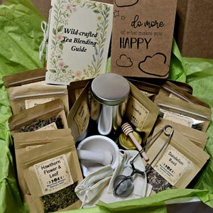Może przedstawiać: Zestaw do mieszania herbaty zawierający przewodnik Wild-crafted Tea Blending Guide, brązowy notatnik z napisem do more of what makes you HAPPY, kilka torebek z papieru kraft z suszonymi ziołami, takimi jak Hawthorn Flower & Leaf i Dandelion Root, metalową puszkę, moździerz z tłuczkiem, drewnianą łyżeczkę do miodu oraz metalowy zaparzacz do herbaty.
