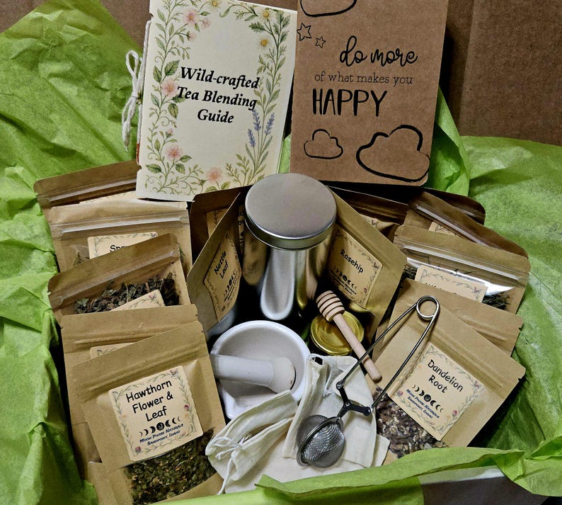 Może przedstawiać: Zestaw do mieszania herbaty zawierający przewodnik Wild-crafted Tea Blending Guide, brązowy notatnik z napisem do more of what makes you HAPPY, kilka torebek z papieru kraft z suszonymi ziołami, takimi jak Hawthorn Flower & Leaf i Dandelion Root, metalową puszkę, moździerz z tłuczkiem, drewnianą łyżeczkę do miodu oraz metalowy zaparzacz do herbaty.