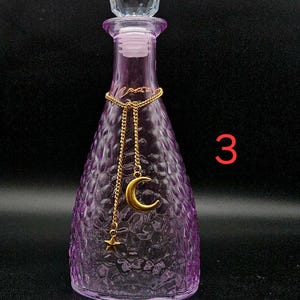 May include: A purple glass bottle with a textured, dimpled surface and a clear, faceted crystal-style stopper. A gold-toned chain is draped around the neck, featuring a hanging gold crescent moon charm and a small gold star charm. The number 3 is visible.