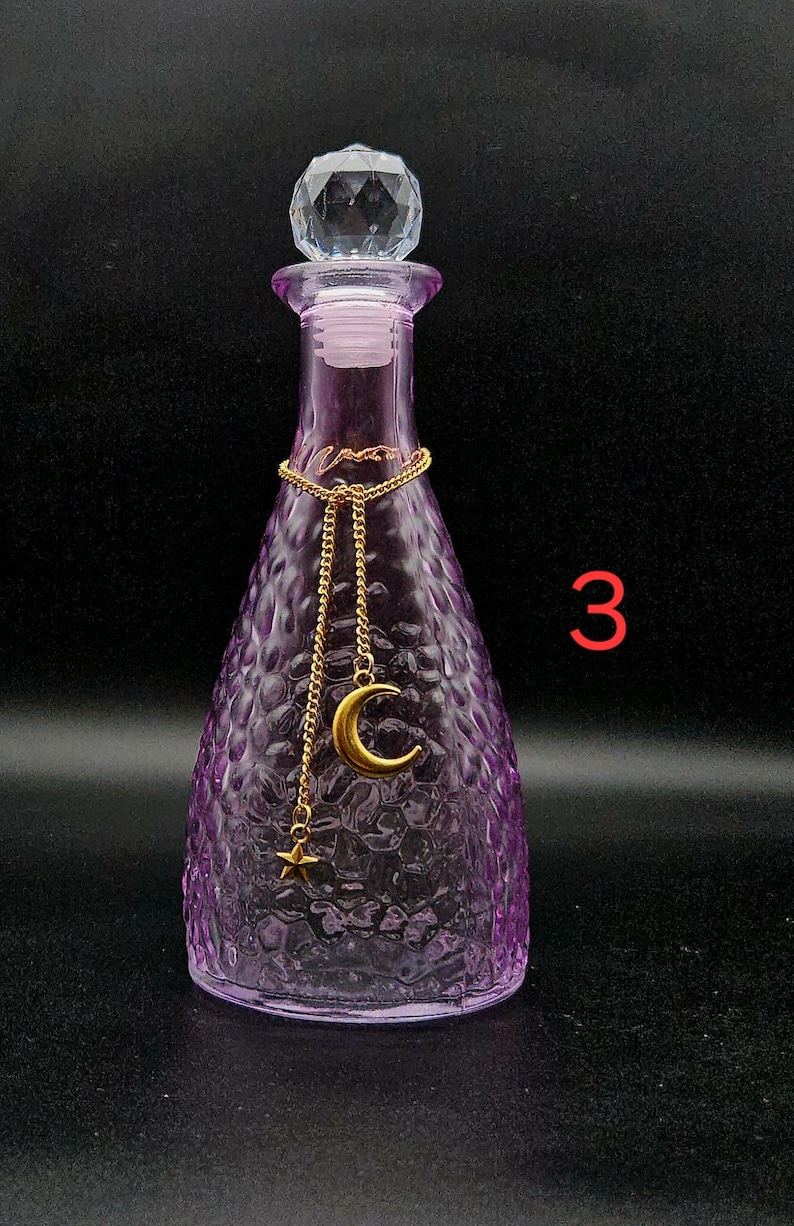 May include: A purple glass bottle with a textured, dimpled surface and a clear, faceted crystal-style stopper. A gold-toned chain is draped around the neck, featuring a hanging gold crescent moon charm and a small gold star charm. The number 3 is visible.