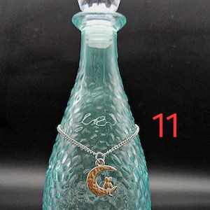May include: A light blue glass bottle with a textured surface and a clear faceted stopper. A silver-toned chain necklace with a crescent moon and cat pendant is draped around the neck of the bottle. The number 11 is printed in red on the right.
