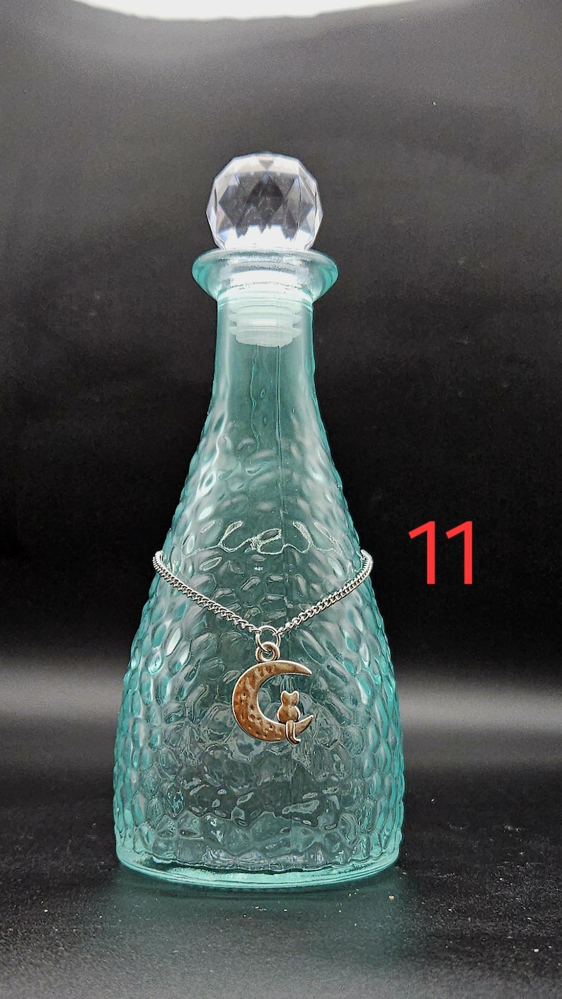 May include: A light blue glass bottle with a textured surface and a clear faceted stopper. A silver-toned chain necklace with a crescent moon and cat pendant is draped around the neck of the bottle. The number 11 is printed in red on the right.