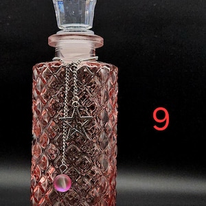 May include: A rectangular pink glass bottle with a diamond-patterned texture, topped with a clear faceted stopper. A silver-toned chain hangs from the neck, featuring a star-shaped charm and a small, round, translucent pink bead. The number 9 is visible.