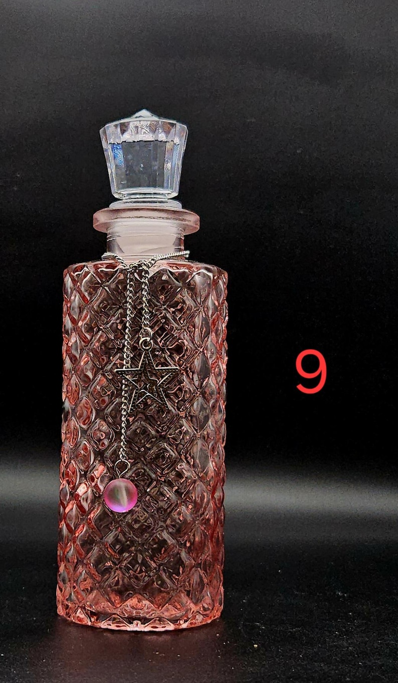 May include: A rectangular pink glass bottle with a diamond-patterned texture, topped with a clear faceted stopper. A silver-toned chain hangs from the neck, featuring a star-shaped charm and a small, round, translucent pink bead. The number 9 is visible.