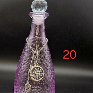 May include: A purple glass bottle with a textured surface and a clear faceted crystal stopper. A silver-toned chain hangs around the neck, featuring a circular sun and moon pendant and a small leaf charm. The number 20 is printed in red on the right.