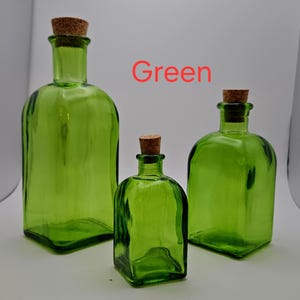 May include: Three green glass bottles with cork stoppers. The bottles are different sizes and have a square base. The word "Green" is written in red. The bottles are a vibrant green colour.