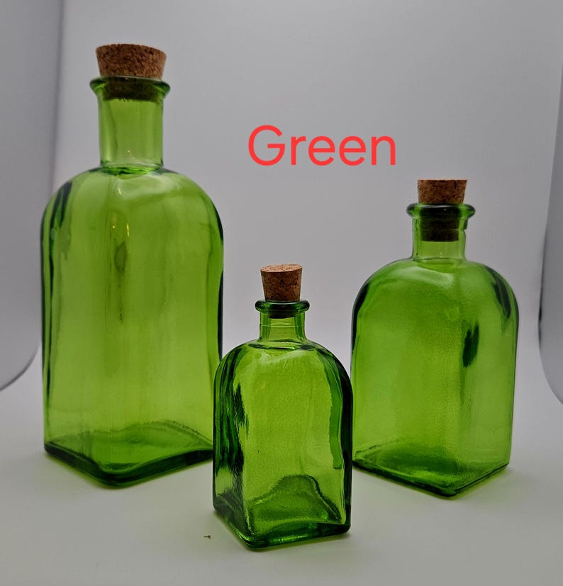 May include: Three green glass bottles with cork stoppers. The bottles are different sizes and have a square base. The word "Green" is written in red. The bottles are a vibrant green colour.