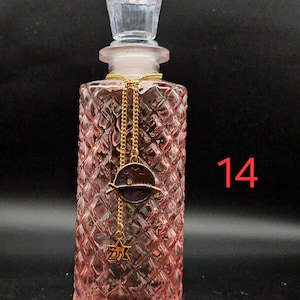 May include: A pink glass perfume bottle with a diamond-cut texture and a clear faceted stopper. A gold-toned chain hangs around the neck, featuring a circular charm with a cat silhouette and a small star-shaped pendant. The number 14 is visible.