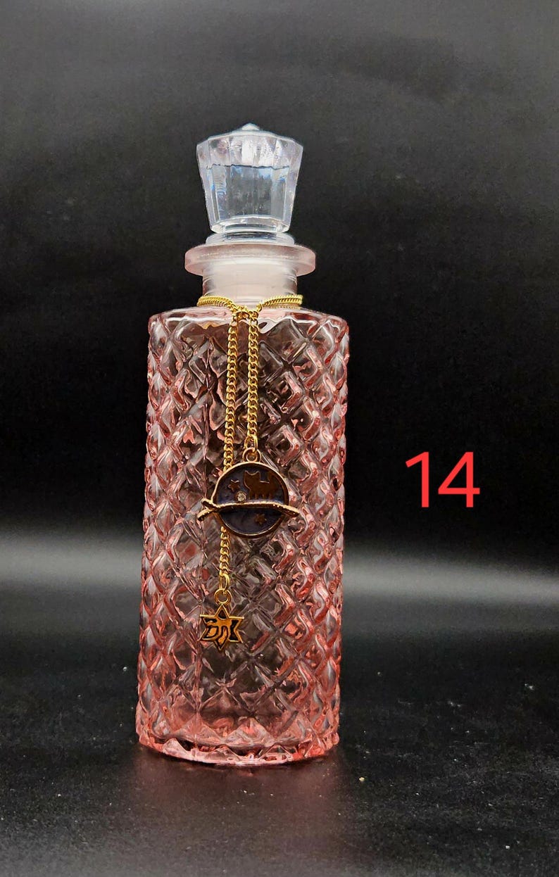 May include: A pink glass perfume bottle with a diamond-cut texture and a clear faceted stopper. A gold-toned chain hangs around the neck, featuring a circular charm with a cat silhouette and a small star-shaped pendant. The number 14 is visible.