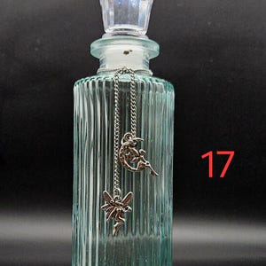 May include: A light green, vertically ribbed glass bottle with a clear, faceted stopper. A silver-toned chain hangs from the neck, featuring two charms: one of a fairy sitting on a crescent moon and another of a flying fairy. The number 17 is visible.