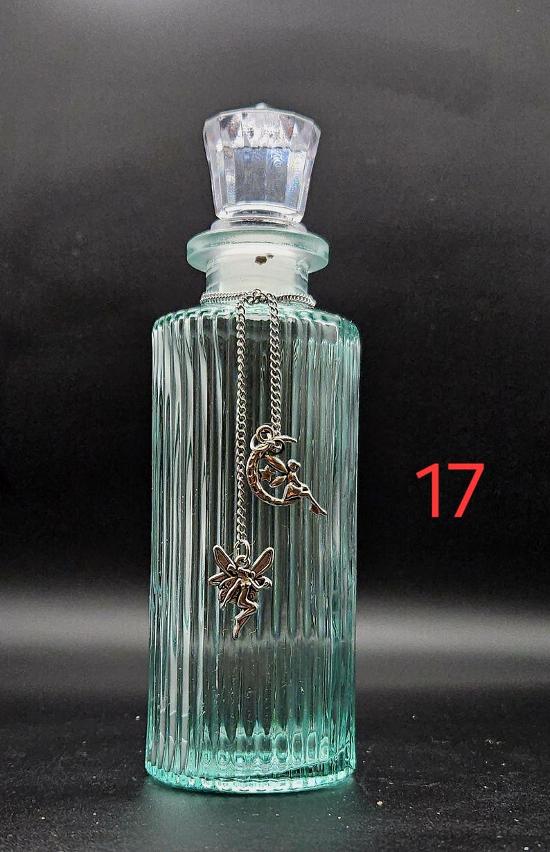 May include: A light green, vertically ribbed glass bottle with a clear, faceted stopper. A silver-toned chain hangs from the neck, featuring two charms: one of a fairy sitting on a crescent moon and another of a flying fairy. The number 17 is visible.
