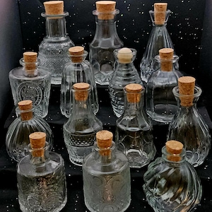 May include: A collection of thirteen clear glass bottles of varying shapes and sizes, each topped with a light brown cork stopper. The bottles feature diverse textures, including smooth, ribbed, diamond-patterned, and embossed glass designs.