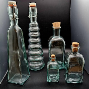 May include: Collection of clear glass bottles with various shapes and sizes. Some bottles have cork stoppers, while others have hinged closures with rubber seals. The bottles are arranged on a dark surface, showcasing their transparent design.