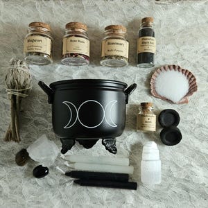 May include: A black cauldron with a white triple moon symbol, surrounded by glass jars labeled Mugwort, Rose Buds, Rosemary, Hawaiian Black Salt, and Frankincense & Myrrh. Includes a sage bundle, white salt in a shell, two charcoal discs, four candles, and crystals.