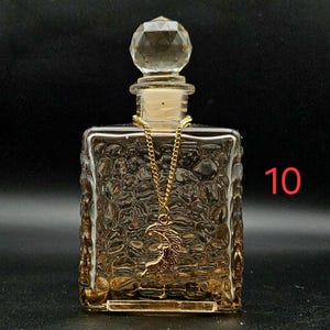 May include: A square glass bottle with a textured, pebbled surface and a clear, faceted, spherical stopper. A gold-toned chain is draped around the neck of the bottle, featuring a crescent moon pendant. The number 10 is printed in red on the right.