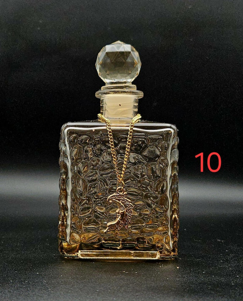 May include: A square glass bottle with a textured, pebbled surface and a clear, faceted, spherical stopper. A gold-toned chain is draped around the neck of the bottle, featuring a crescent moon pendant. The number 10 is printed in red on the right.