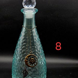 May include: A light blue glass bottle with a textured, dimpled surface and a clear, faceted crystal-like stopper. A thin bronze-toned chain hangs around the neck, featuring a circular sun and moon pendant and a small leaf charm. The number 8 is visible.