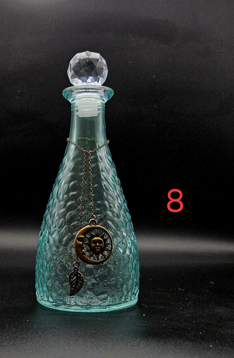 May include: A light blue glass bottle with a textured, dimpled surface and a clear, faceted crystal-like stopper. A thin bronze-toned chain hangs around the neck, featuring a circular sun and moon pendant and a small leaf charm. The number 8 is visible.