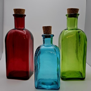 May include: Three coloured glass bottles with cork stoppers. The bottles are in shades of red, blue, and green. The bottles have a rectangular shape with rounded edges. The corks are a light brown colour.