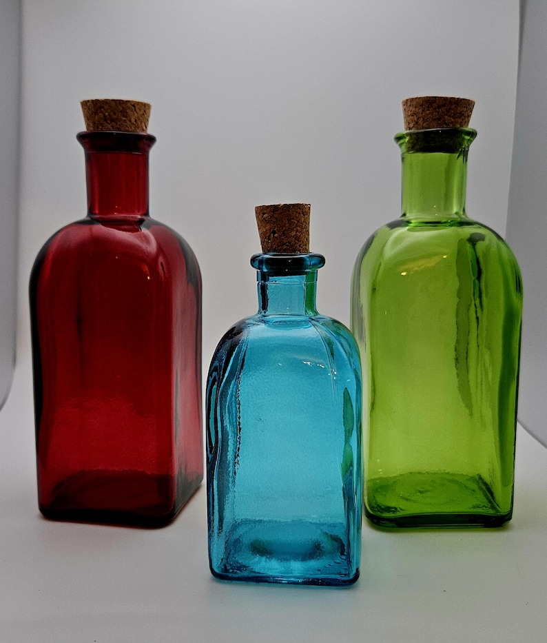 May include: Three coloured glass bottles with cork stoppers. The bottles are in shades of red, blue, and green. The bottles have a rectangular shape with rounded edges. The corks are a light brown colour.