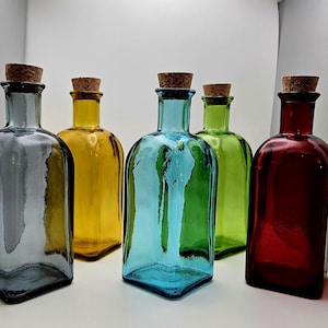 May include: Five coloured glass bottles with cork stoppers. The bottles are in shades of grey, yellow, blue, green, and red. The bottles have a rectangular shape with rounded shoulders.