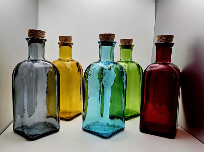 May include: Five coloured glass bottles with cork stoppers. The bottles are in shades of grey, yellow, blue, green, and red. The bottles have a rectangular shape with rounded shoulders.