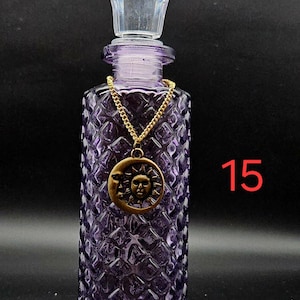 May include: A purple glass bottle with a diamond-patterned texture and a clear faceted stopper. A gold-toned chain hangs around the neck, featuring a circular pendant with a sun and moon design. The number 15 is printed in red on the right side.