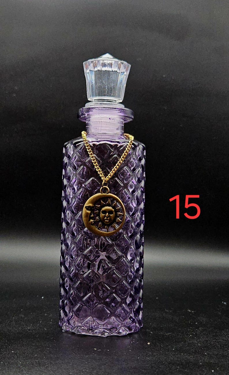 May include: A purple glass bottle with a diamond-patterned texture and a clear faceted stopper. A gold-toned chain hangs around the neck, featuring a circular pendant with a sun and moon design. The number 15 is printed in red on the right side.