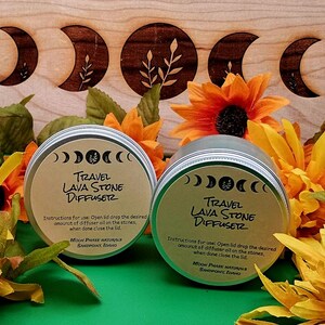 May include: Two silver travel lava stone diffusers with the text "Travel Lava Stone Diffuser" and instructions. The diffusers are surrounded by orange and yellow flowers, with a wooden background.